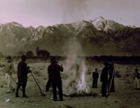 1943 Burning leaves, autumn dawn, Manzanar Relocation Center, California / photograph by Ansel Adams. SUMMARY: Group of men and boys standing around a small brush fire holding shovels, a pitchfork stands in the right foreground, mountains in background. V