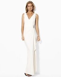 The elegant floor-grazing gown is rendered in a sleeveless silhouette with a crossover V-neckline secured with an encrusted broach and finished with a graceful ruffle the length of the skirt. The elegant floor-grazing gown is rendered in a sleeveless silhouette with a crossover V-neckline secured with an encrusted broach and finished with a graceful ruffle the length of the skirt.